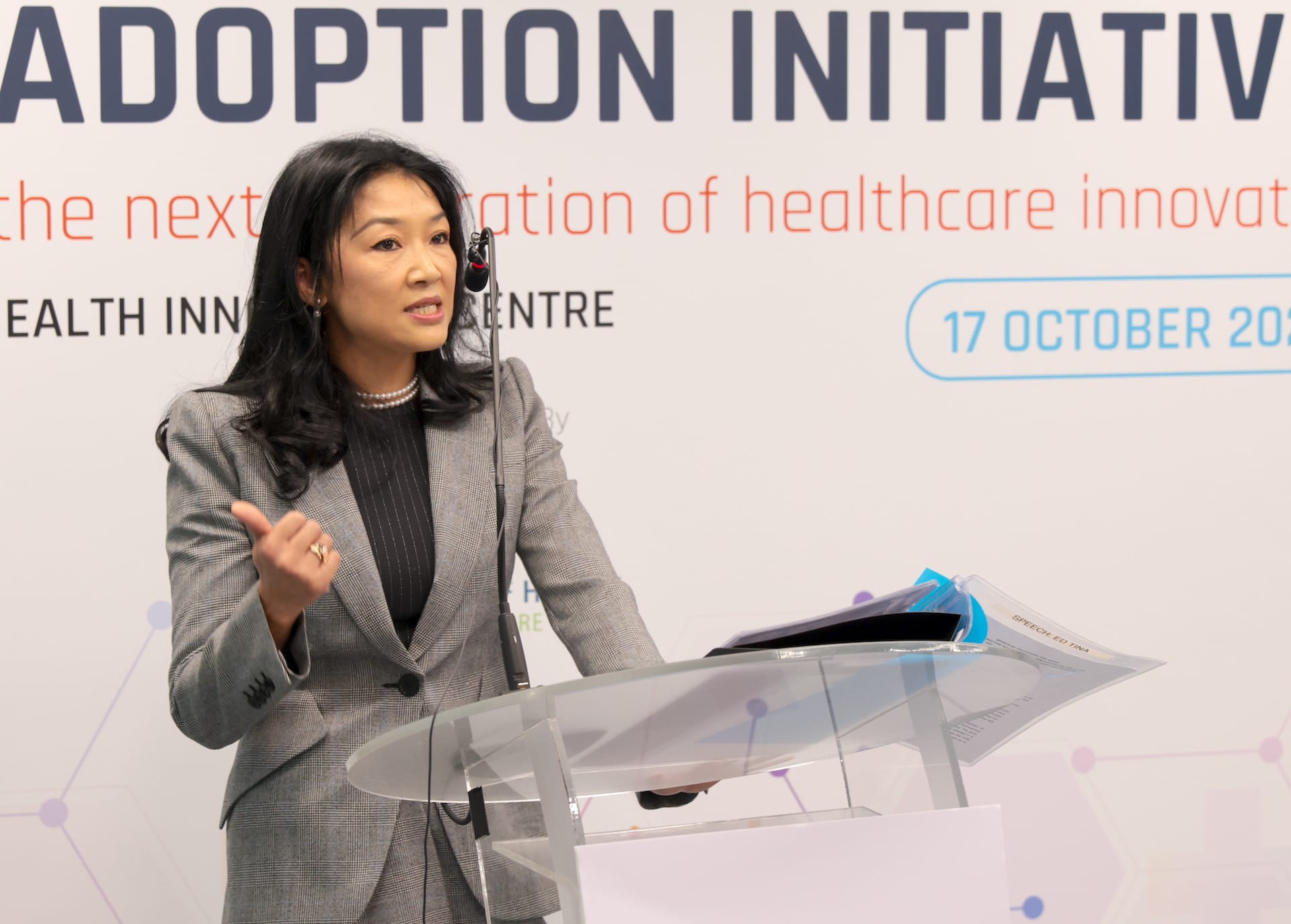 A woman in a grey suit speaks at a podium with an event banner in the background mentioning healthcare innovation.